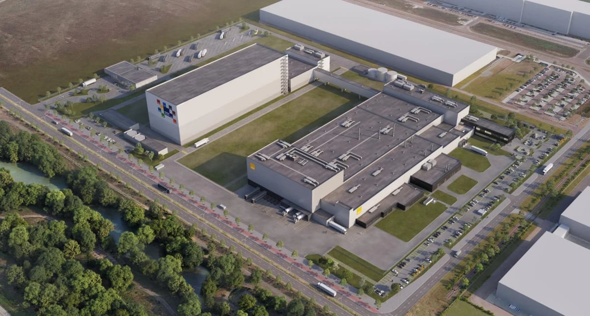 Aerial view of industrial manufacturing facility with multiple large warehouses, parking lots, and green spaces adjacent to a highway.